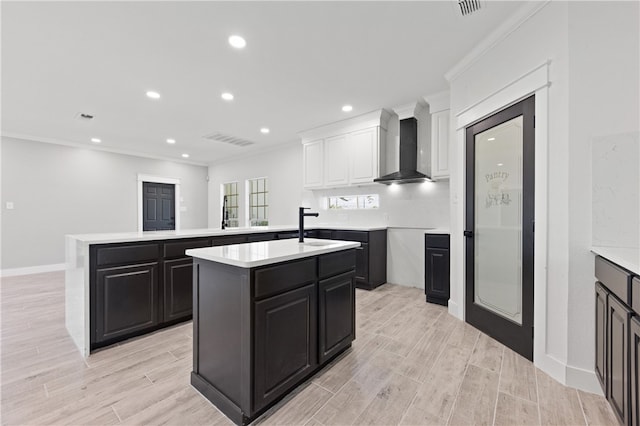 kitchen with an island with sink, wall chimney range hood, light hardwood / wood-style flooring, and ornamental molding