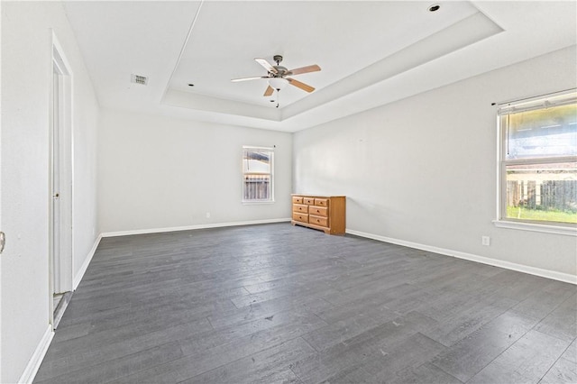 spare room featuring dark wood-type flooring, a ceiling fan, a raised ceiling, and a healthy amount of sunlight