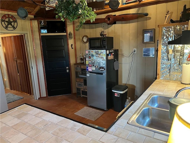 kitchen featuring wooden walls, tile counters, freestanding refrigerator, black microwave, and a sink