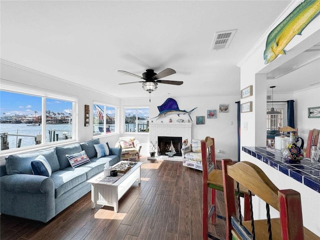 living area with crown molding, a fireplace with raised hearth, a ceiling fan, dark wood-type flooring, and visible vents