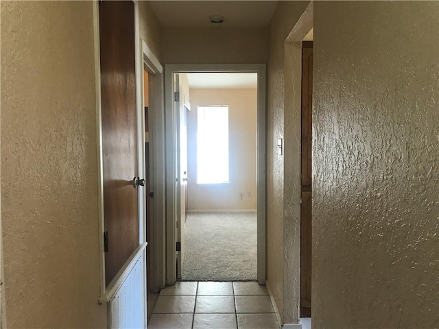 hallway featuring light carpet, light tile patterned floors, baseboards, and a textured wall