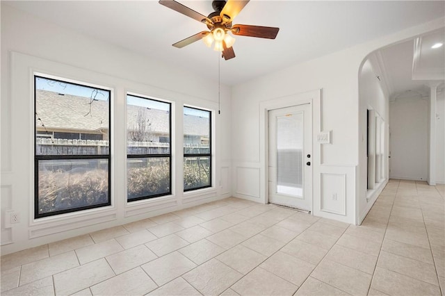 spare room featuring light tile patterned floors, arched walkways, a decorative wall, a ceiling fan, and wainscoting