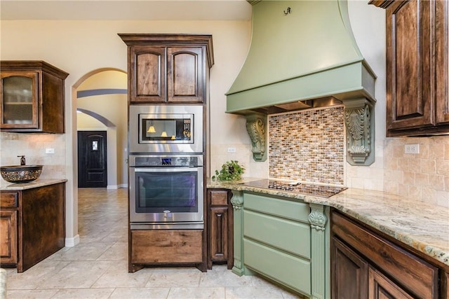 kitchen with light stone counters, custom exhaust hood, arched walkways, dark brown cabinetry, and appliances with stainless steel finishes