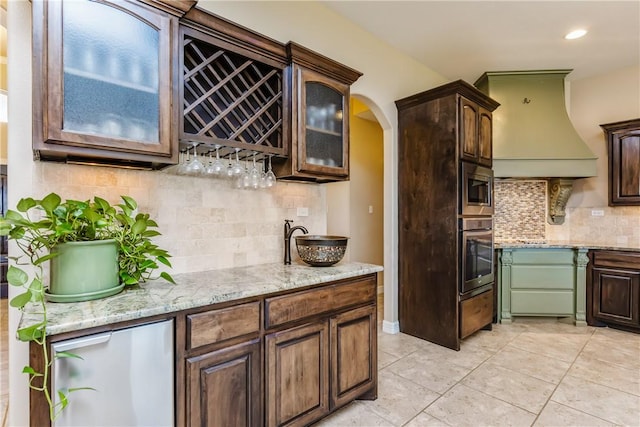 kitchen with stainless steel appliances, arched walkways, glass insert cabinets, light stone countertops, and custom exhaust hood