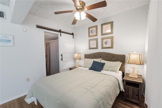 bedroom featuring a barn door, dark wood-type flooring, a ceiling fan, a textured ceiling, and baseboards