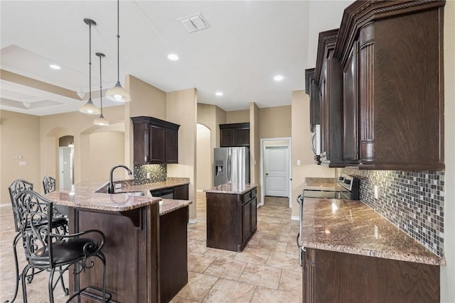 kitchen with dark brown cabinetry, light stone counters, arched walkways, stainless steel fridge, and a sink