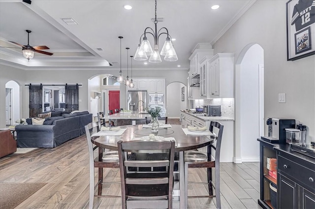 dining space featuring a tray ceiling, a barn door, ceiling fan with notable chandelier, and ornamental molding