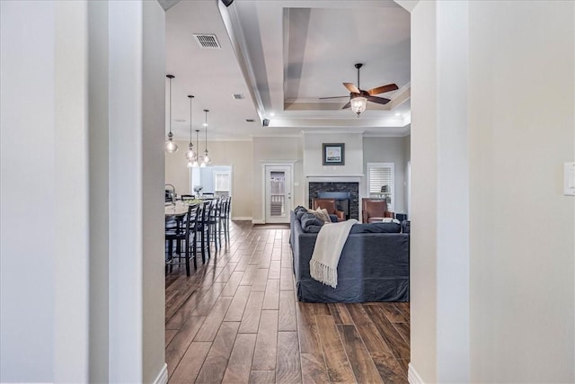 living room with ceiling fan, a stone fireplace, crown molding, and a tray ceiling