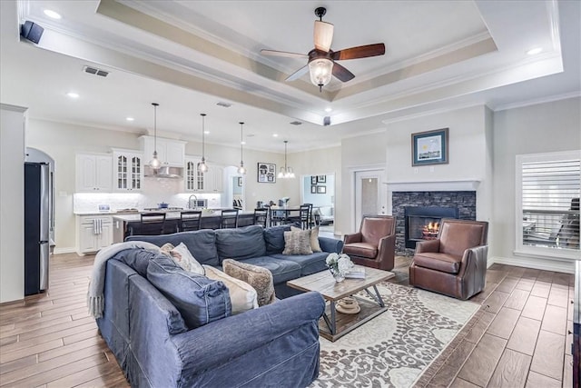 living room with ceiling fan, a stone fireplace, a raised ceiling, and ornamental molding