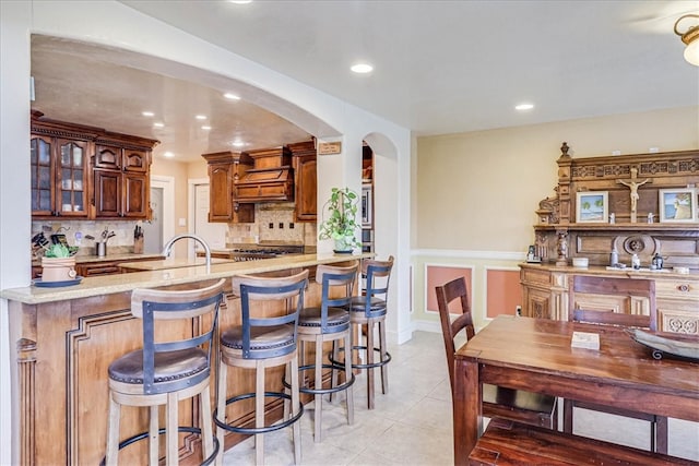 kitchen featuring kitchen peninsula, light tile patterned flooring, backsplash, and premium range hood