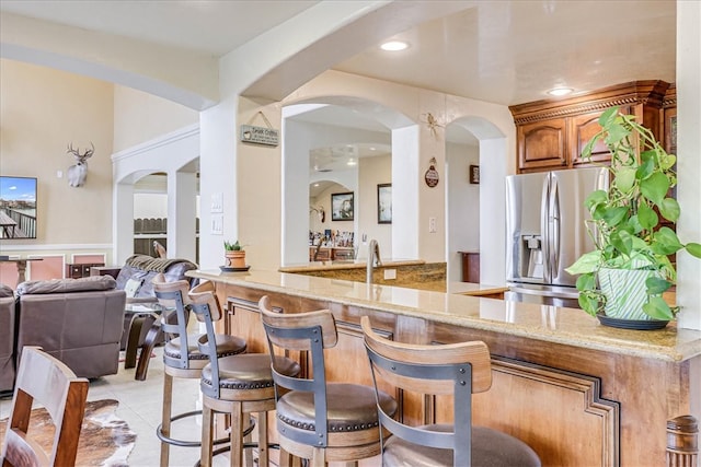 bar featuring stainless steel refrigerator with ice dispenser, light tile patterned floors, and light stone countertops