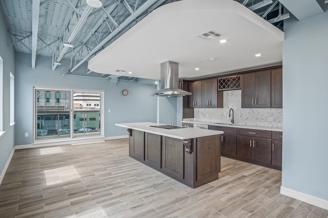 kitchen with light wood-type flooring, dark brown cabinets, sink, wall chimney exhaust hood, and a kitchen island with sink