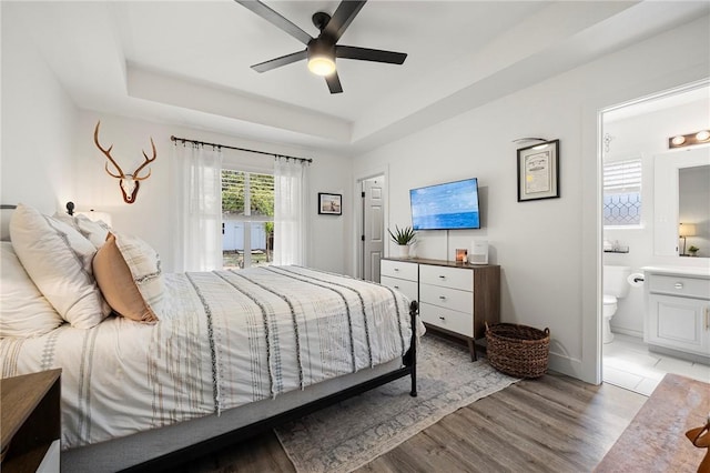 bedroom featuring a ceiling fan, light wood-type flooring, baseboards, ensuite bathroom, and a tray ceiling