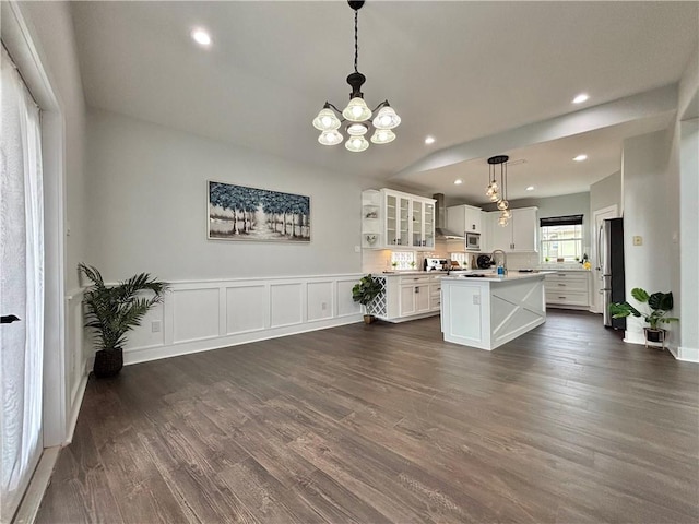 kitchen featuring wall chimney range hood, hanging light fixtures, dark wood-type flooring, and an island with sink