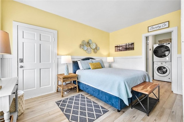 bedroom featuring light wood-style flooring, a decorative wall, stacked washer / drying machine, and wainscoting