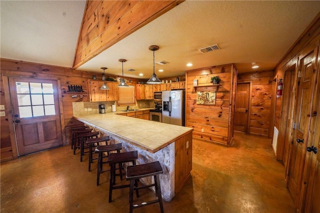 kitchen with kitchen peninsula, hanging light fixtures, stainless steel appliances, wooden walls, and a kitchen bar