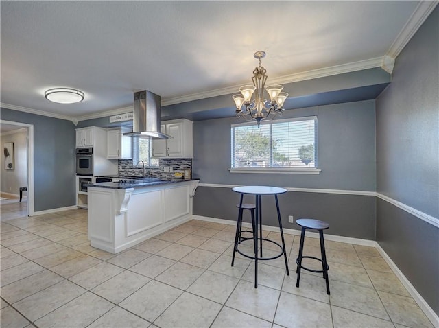 kitchen with wall chimney range hood, a notable chandelier, kitchen peninsula, decorative backsplash, and white cabinets