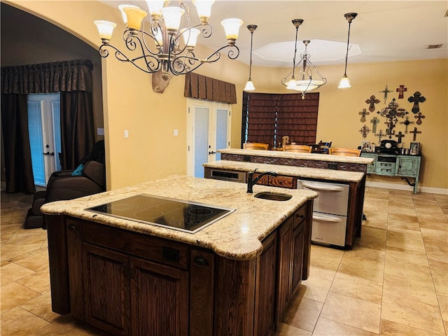 kitchen featuring black electric stovetop, sink, a kitchen island with sink, light stone countertops, and pendant lighting