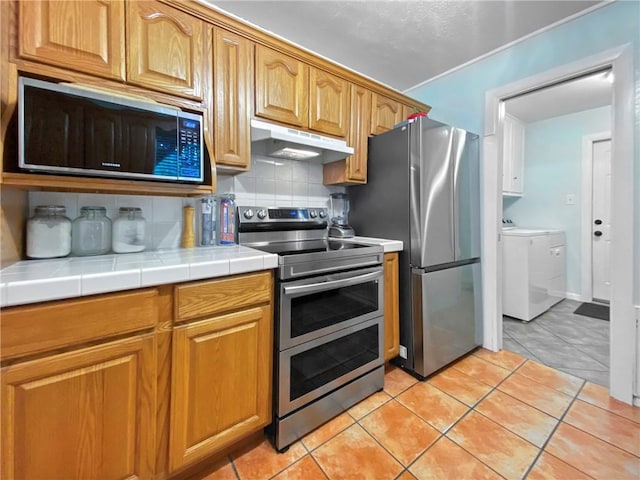 kitchen featuring stainless steel appliances, tile countertops, light tile patterned flooring, decorative backsplash, and washing machine and dryer