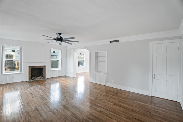 unfurnished living room featuring built in shelves, ornamental molding, hardwood / wood-style flooring, a brick fireplace, and ceiling fan