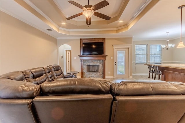 living room with crown molding, a raised ceiling, ceiling fan with notable chandelier, and a stone fireplace