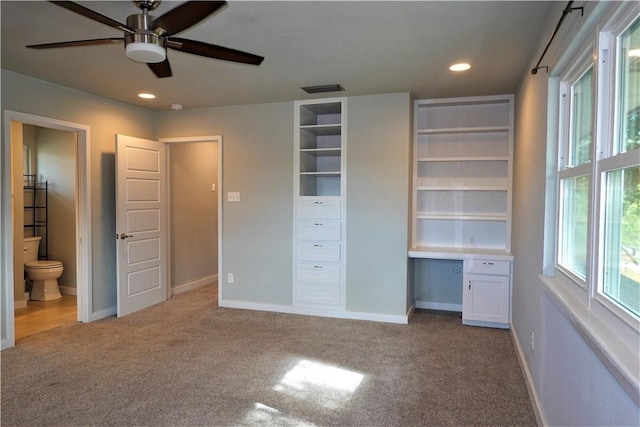 unfurnished bedroom featuring light carpet, built in desk, ensuite bathroom, and ceiling fan