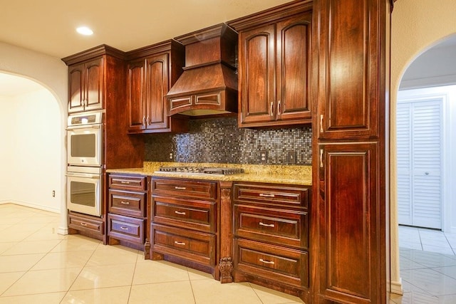 kitchen with stainless steel gas stovetop, light stone counters, decorative backsplash, custom exhaust hood, and white double oven