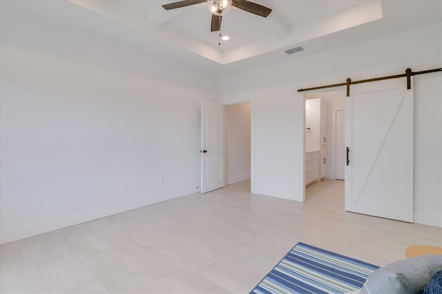 unfurnished bedroom featuring connected bathroom, ceiling fan, a barn door, a tray ceiling, and light tile patterned floors