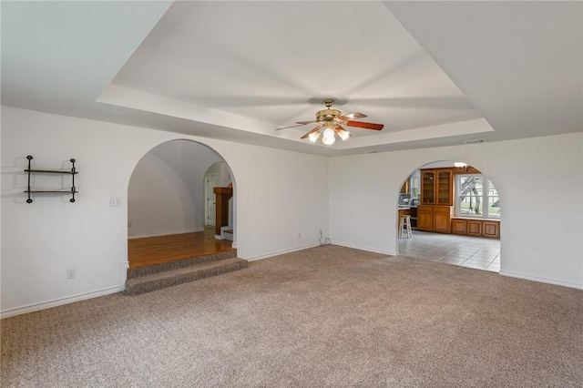 unfurnished room featuring arched walkways, a raised ceiling, light carpet, ceiling fan, and baseboards