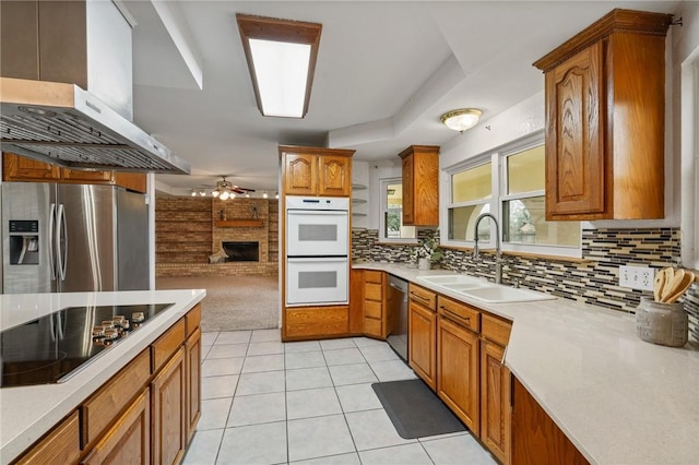 kitchen with a brick fireplace, stainless steel appliances, light countertops, wall chimney range hood, and a sink