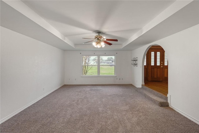 empty room with baseboards, arched walkways, a raised ceiling, a ceiling fan, and light colored carpet