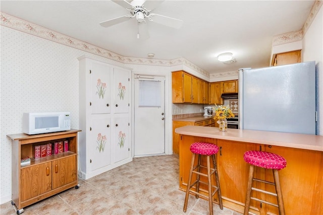 kitchen with brown cabinets, wallpapered walls, freestanding refrigerator, light countertops, and white microwave