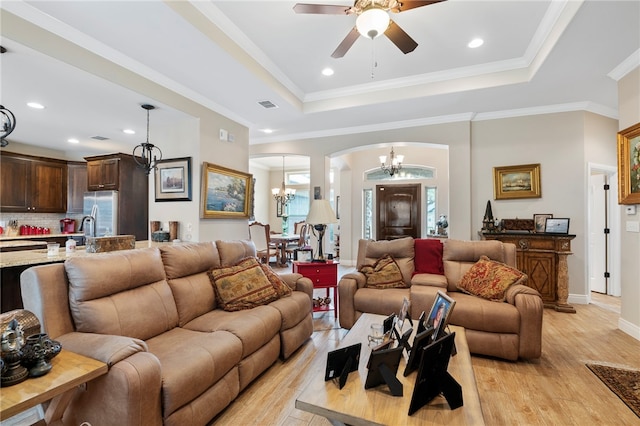 living room with ceiling fan with notable chandelier, a raised ceiling, ornamental molding, and light hardwood / wood-style flooring