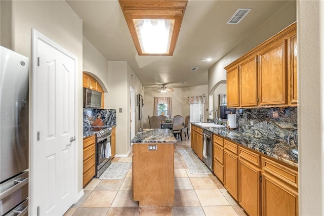 kitchen featuring arched walkways, a kitchen island, appliances with stainless steel finishes, dark stone countertops, and a sink