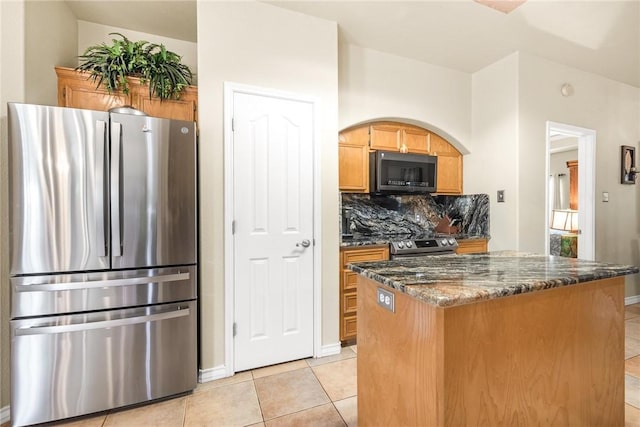 kitchen with tasteful backsplash, appliances with stainless steel finishes, brown cabinetry, light tile patterned flooring, and dark stone countertops