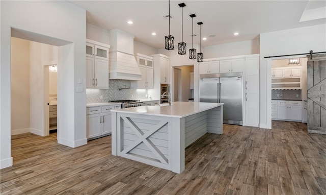 kitchen featuring built in appliances, a barn door, wood finished floors, custom exhaust hood, and white cabinets