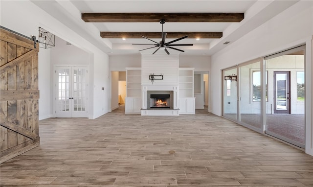 unfurnished living room featuring a large fireplace, a barn door, beam ceiling, light wood-style flooring, and french doors