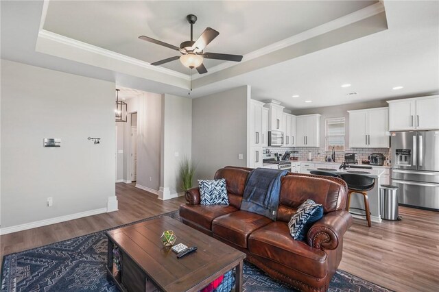 living room featuring ceiling fan, baseboards, light wood-type flooring, a tray ceiling, and ornamental molding