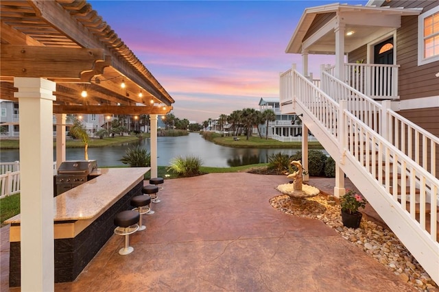 patio terrace at dusk with an outdoor kitchen, a water view, area for grilling, and exterior bar