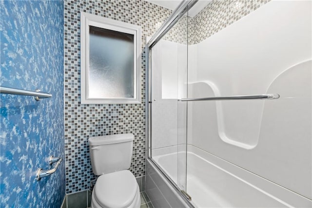 bathroom featuring toilet, combined bath / shower with glass door, and tile walls