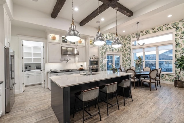 kitchen with wallpapered walls, under cabinet range hood, beam ceiling, appliances with stainless steel finishes, and a sink