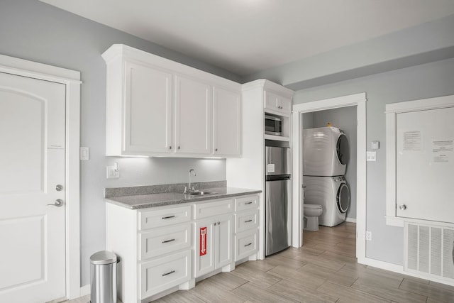 kitchen with appliances with stainless steel finishes, sink, stacked washer / dryer, white cabinetry, and light hardwood / wood-style flooring