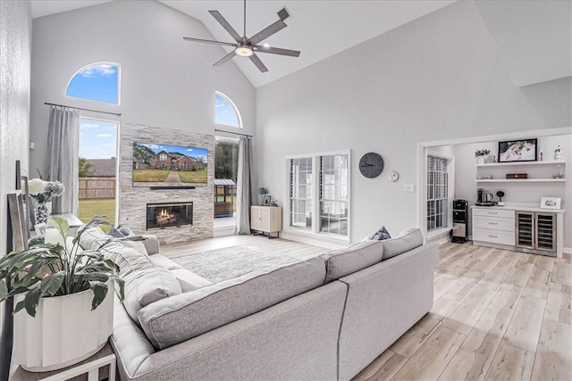 living room featuring high vaulted ceiling, a stone fireplace, ceiling fan, light hardwood / wood-style floors, and beverage cooler