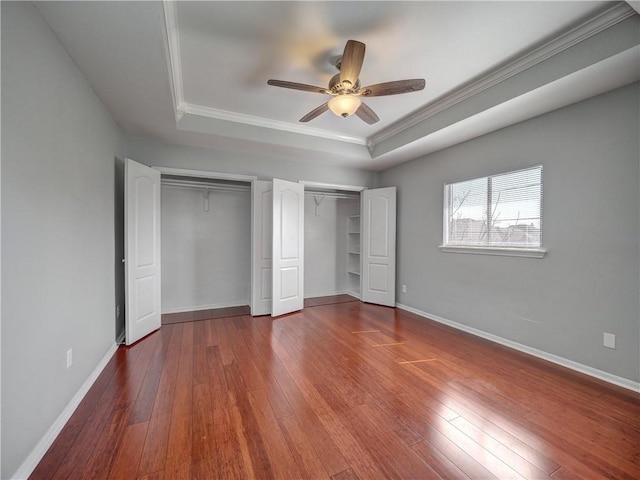 unfurnished bedroom featuring a tray ceiling, hardwood / wood-style flooring, baseboards, and two closets