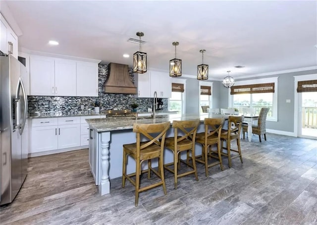 kitchen featuring a center island with sink, white cabinetry, custom range hood, stainless steel refrigerator with ice dispenser, and hanging light fixtures