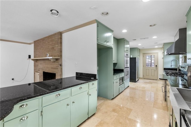 kitchen featuring wall chimney exhaust hood, crown molding, dark stone counters, a fireplace, and green cabinetry