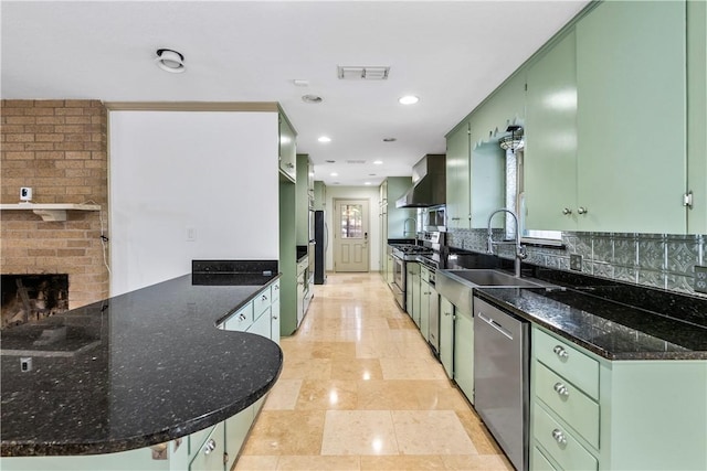 kitchen featuring wall chimney range hood, sink, green cabinetry, appliances with stainless steel finishes, and kitchen peninsula