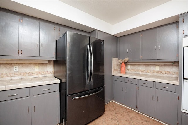 kitchen with light tile patterned floors, tile counters, decorative backsplash, freestanding refrigerator, and gray cabinetry