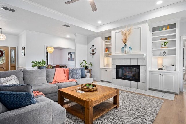 living room featuring a ceiling fan, light wood-type flooring, a tile fireplace, a tray ceiling, and ornamental molding