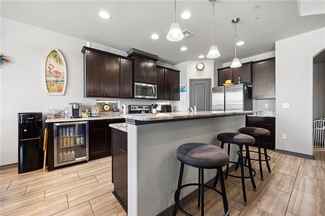 kitchen featuring beverage cooler, an island with sink, dark brown cabinetry, appliances with stainless steel finishes, and a kitchen bar